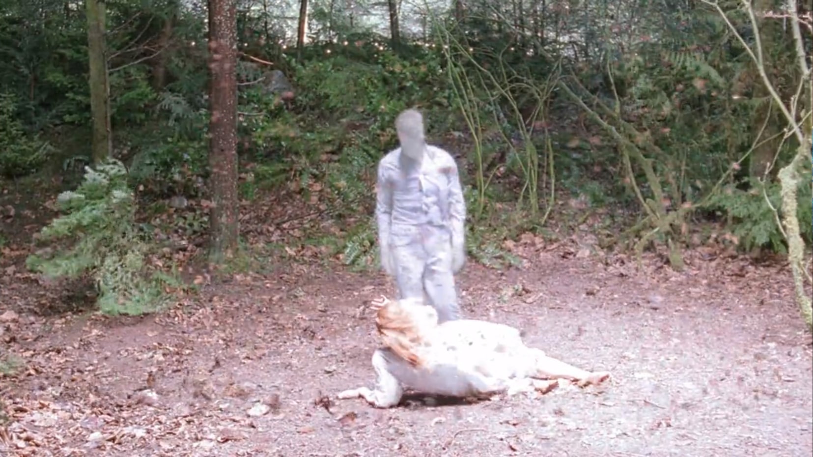 A girl in a white night dress is on the ground in a forest clearing her back to us as she cowers before what appears to be a man made of static. He is wearing a button up shirt, though the color is not clear as his form appears to be washed out. An intense unnatural light shines down on them from above. The wind is cycloning around them, making the leaves fly through the air.