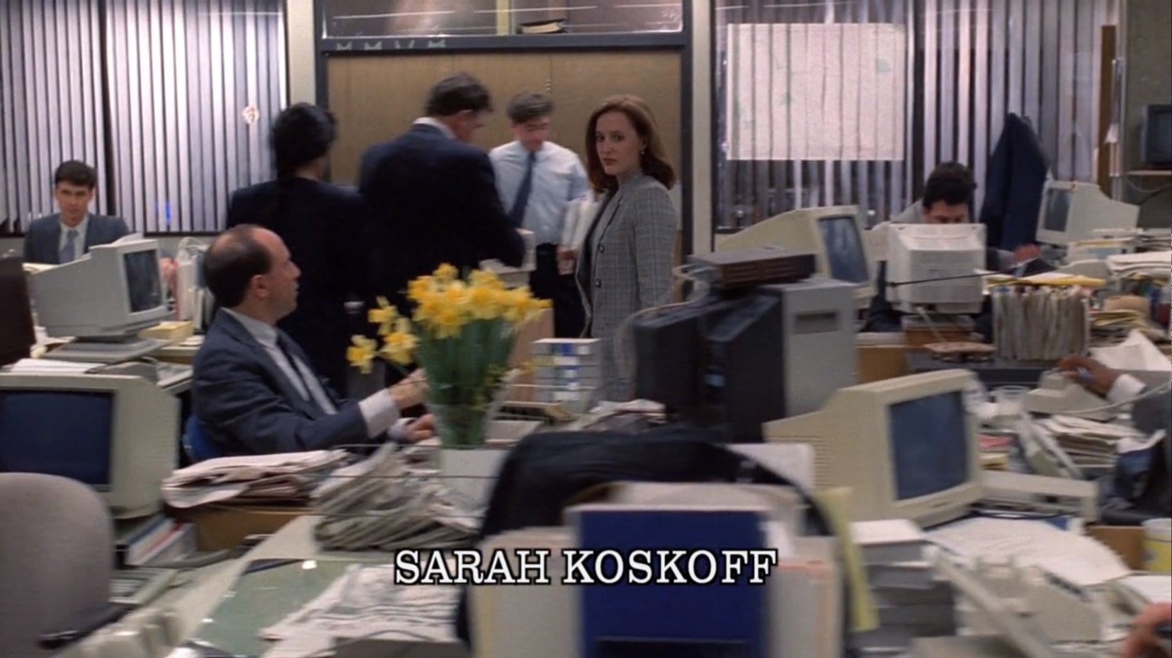 Scully walks briskly through a busy large office area, where numerous cluttered desks can be seen. Some FBI agents are in the background, their faces either blurry or turned from the camera, presumably working. There is one man sitting in the foreground next to a vase filled with yellow buttercups. All of the computer monitors, all big old-school CRTS, are turned off.