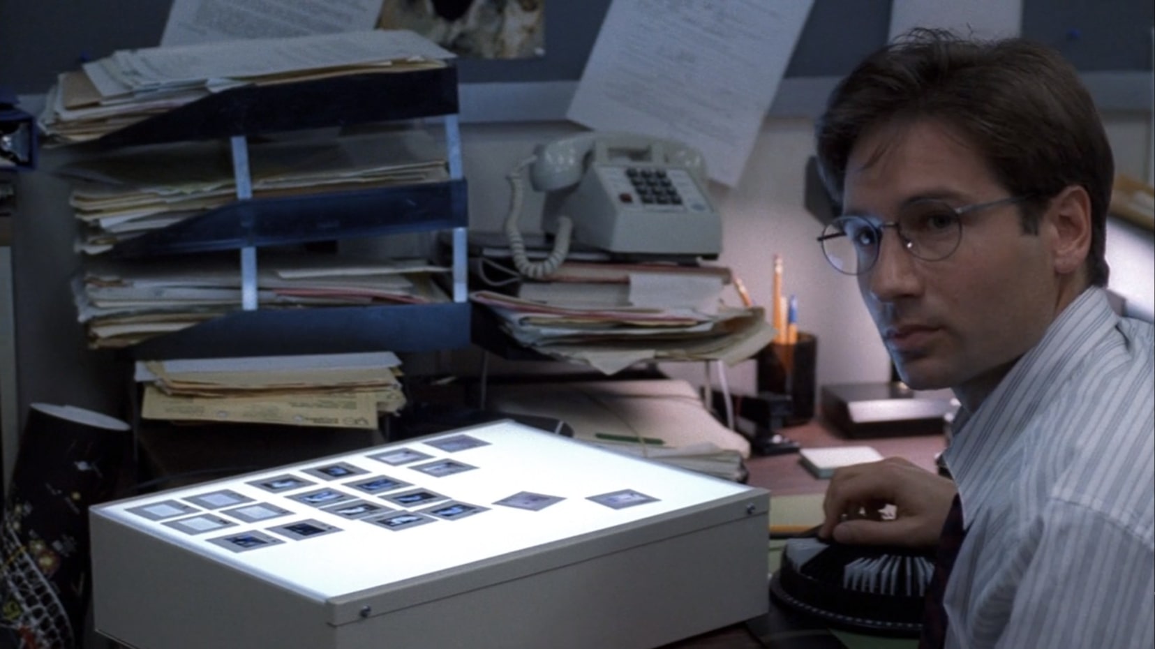 David Duchovny, playing Fox Mulder, looks up from his work in a cluttered office space. He's wearing round glasses and a thin striped button up shirt. On the desk in front of him is a lightbox, where some slides are arranged in rows for examination.