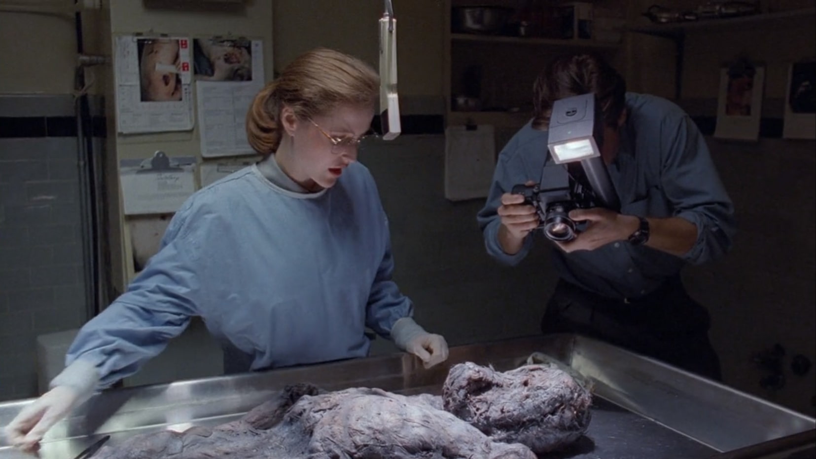 Scully is dressed in a blue scrub with medical gloves pulled on. She's wearing glasses and has her red hair pulled back into a ponytail. She is looking down at a strange disfigured body in advanced decomposition on a metal autopsy table. The face looks unrecognizable. Mulder, to her left, wearing a blue button up long-sleeved shirt with the sleeves rolled up and a dark watch on his wrist, leans in to take a picture with his high quality camera. On the walls behind them are clipboards filled with forms and printouts along with autopsy photos.