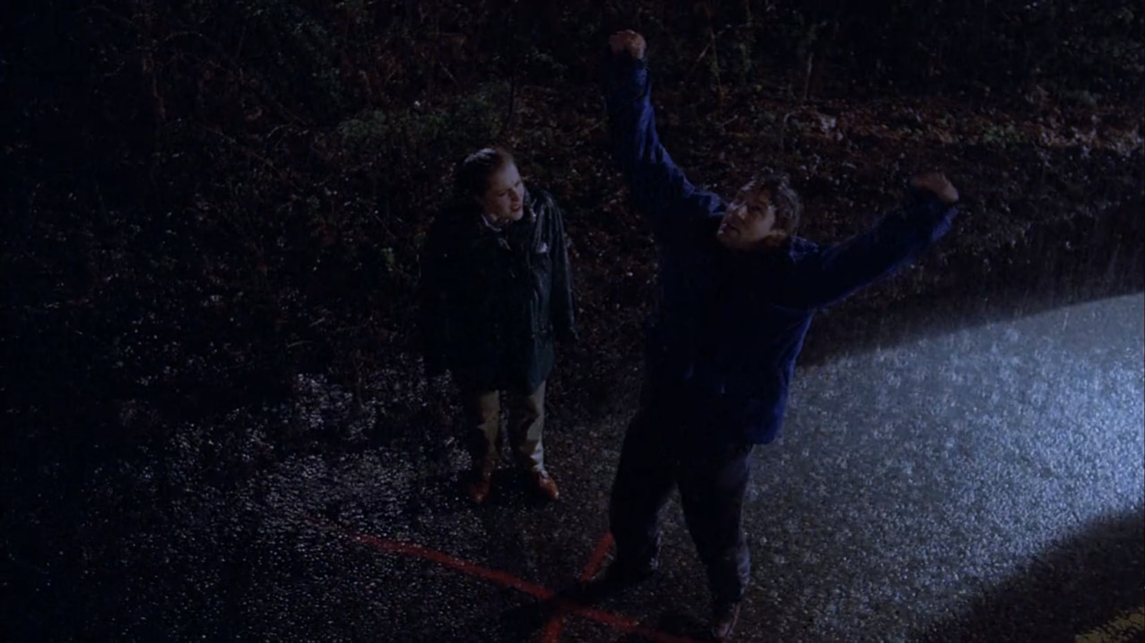 It's nighttime and it's raining. Scully and Mulder are standing on a country road wearing rain coats. At their feet is a red X Mulder had spray-painted earlier that day. He's looking up at the sky, his fists raised in the air, a triumphant expression on his face. Scully, to his right, stares at him like he's crazy. Her hair is pulled back in a ponytail.