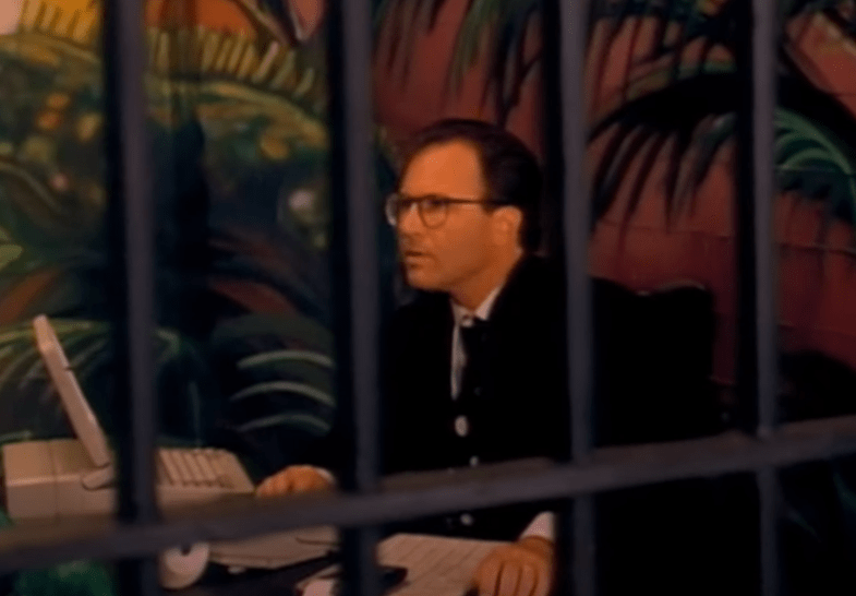A picture of Gary Lee's financial assistant inside the prison cell, sitting at a desk with what looks like a small computer or maybe even a digital typewriter. He's wearing dark glasses and a dark suit with a black tie.