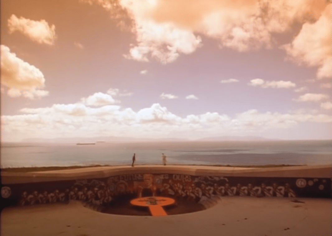 A distant wide shot of Griffin at the graffiti-covered skate park with the ocean visible in the distance.